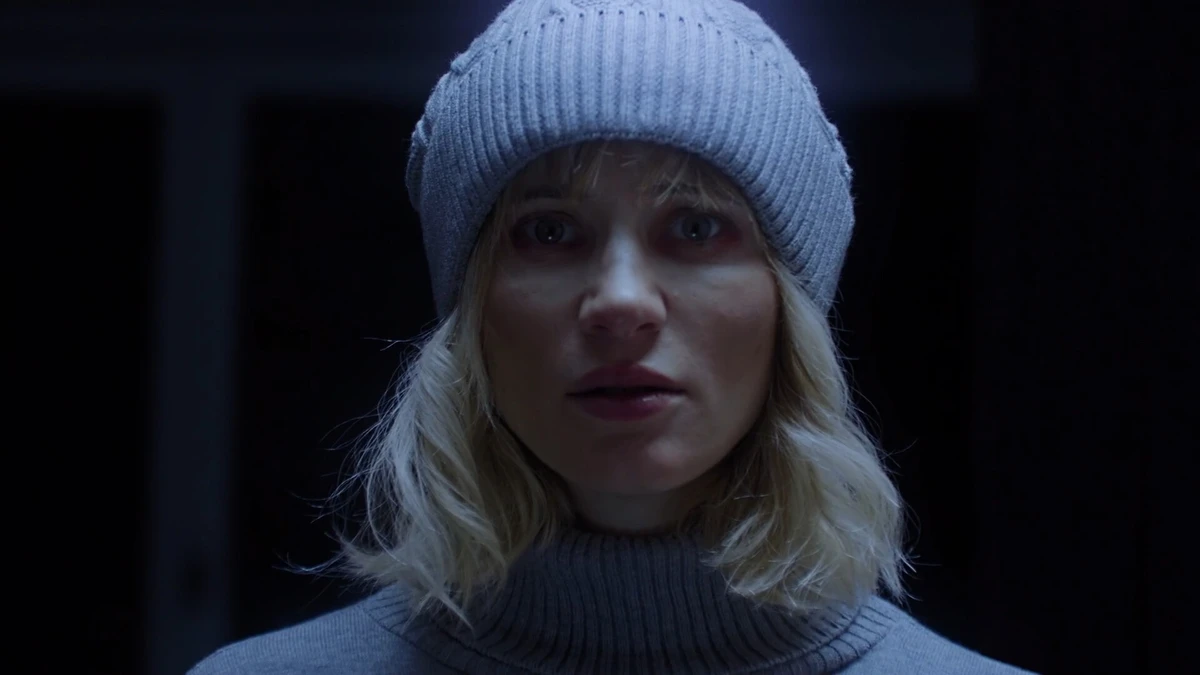 Close-up of a woman wearing a knit hat and turtleneck in a dark horror setting.