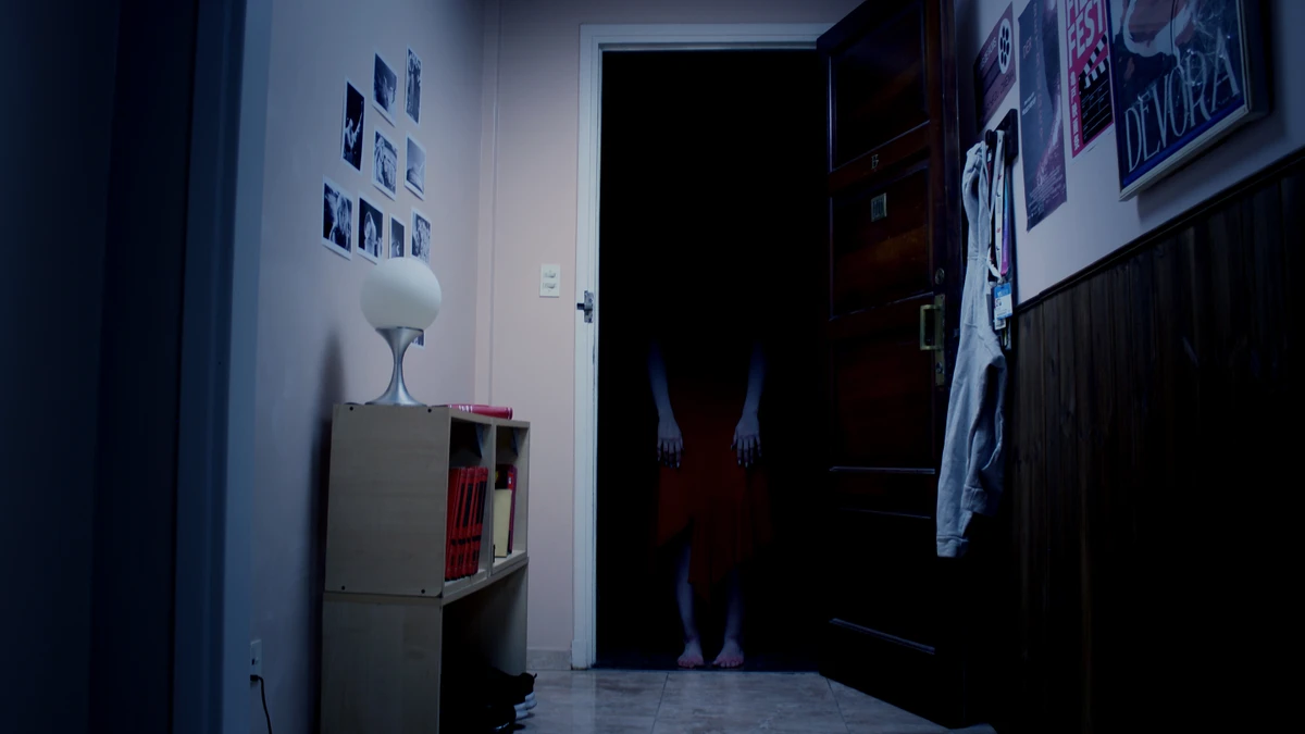 A dark horror-game hallway with an open doorway and a shadowy figure standing inside.